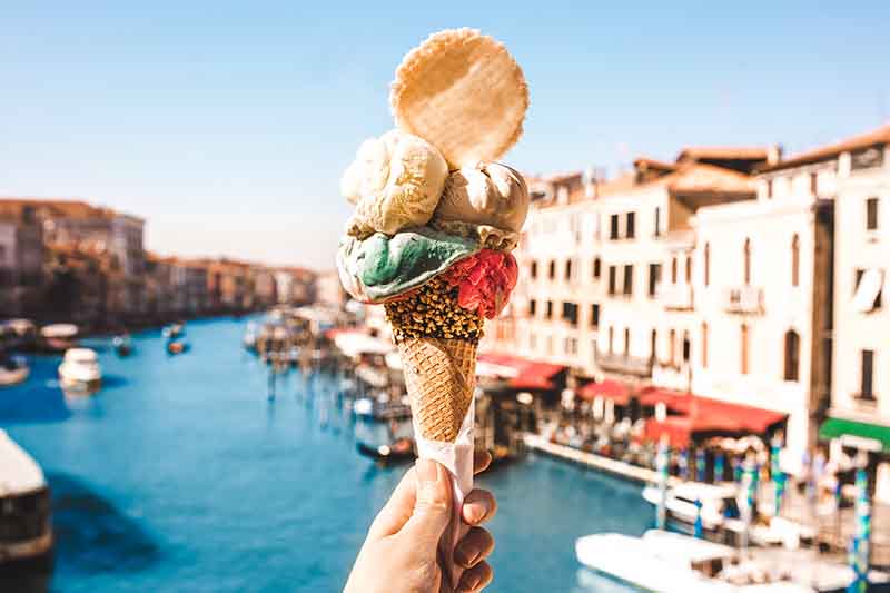 Horizontal image of a hand holding scoops of gelato and a cookie garnish in a cone, with a riverfront background.