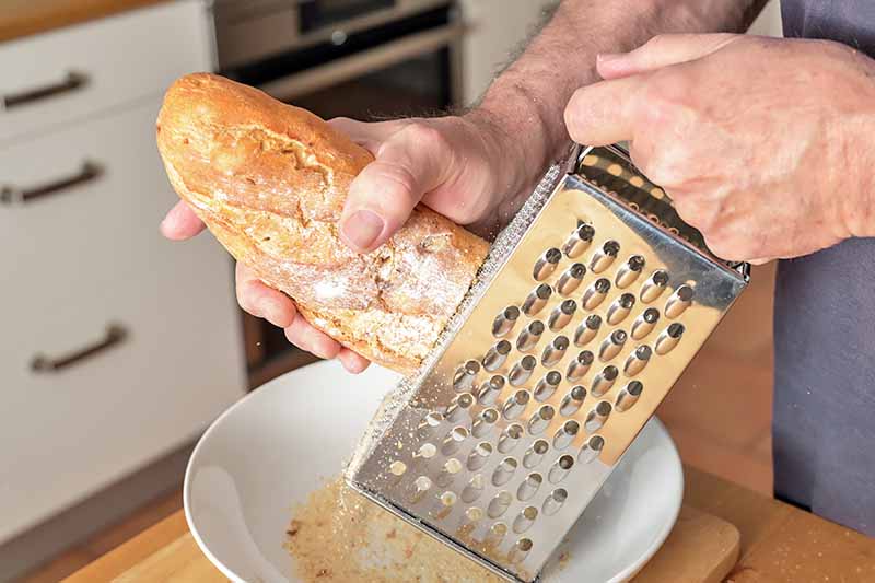 Horizontal image of hands grating a piece of baguette on a box grater over a bowl.