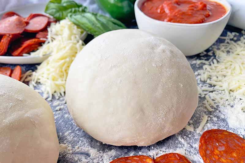Horizontal image of a mound of lightly floured dough next to shredded cheese, basil, pepperoni slices, and a bowl of marinara sauce.