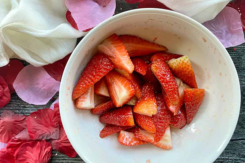 Horizontal image of sliced strawberries in a white bowl next to rose petals.