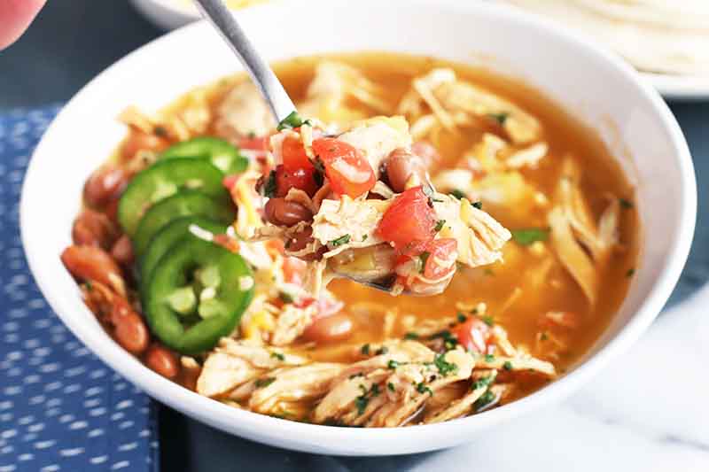 Horizontal image of a spoon holding up some stew with shredded poultry and beans over a bowl of the same recipe, garnished with jalapeno slices on the side.