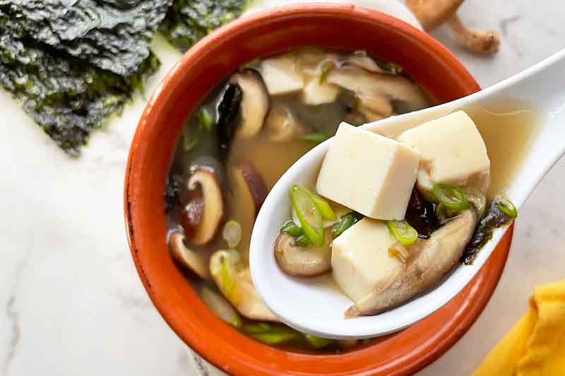 Horizontal image of spoon holding cubes of tofu with scallions and mushrooms over a bowl of broth.