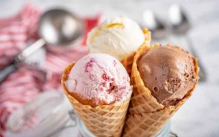 Horizontal image of scoops of frozen dessert in waffle cones in front of a metal scooper on a red towel and spoons.