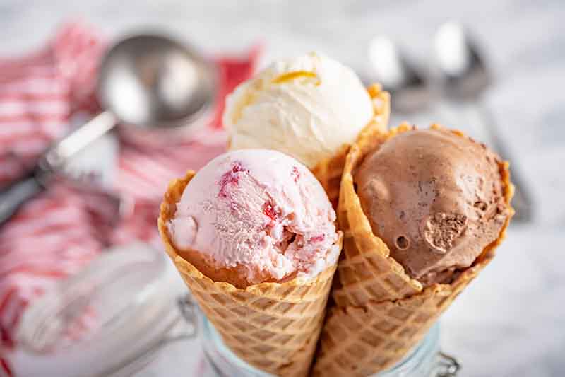 Horizontal image of scoops of frozen dessert in waffle cones in front of a metal scooper on a red towel and spoons.