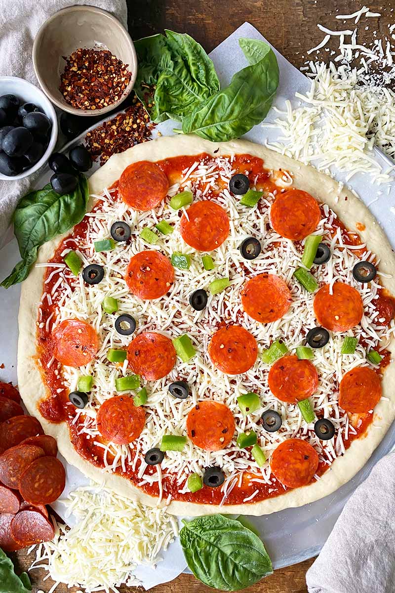 Vertical image of an unbaked pizza with marinara, pepperoni, green peppers, olives, and shredded cheese surrounded by the same toppings.