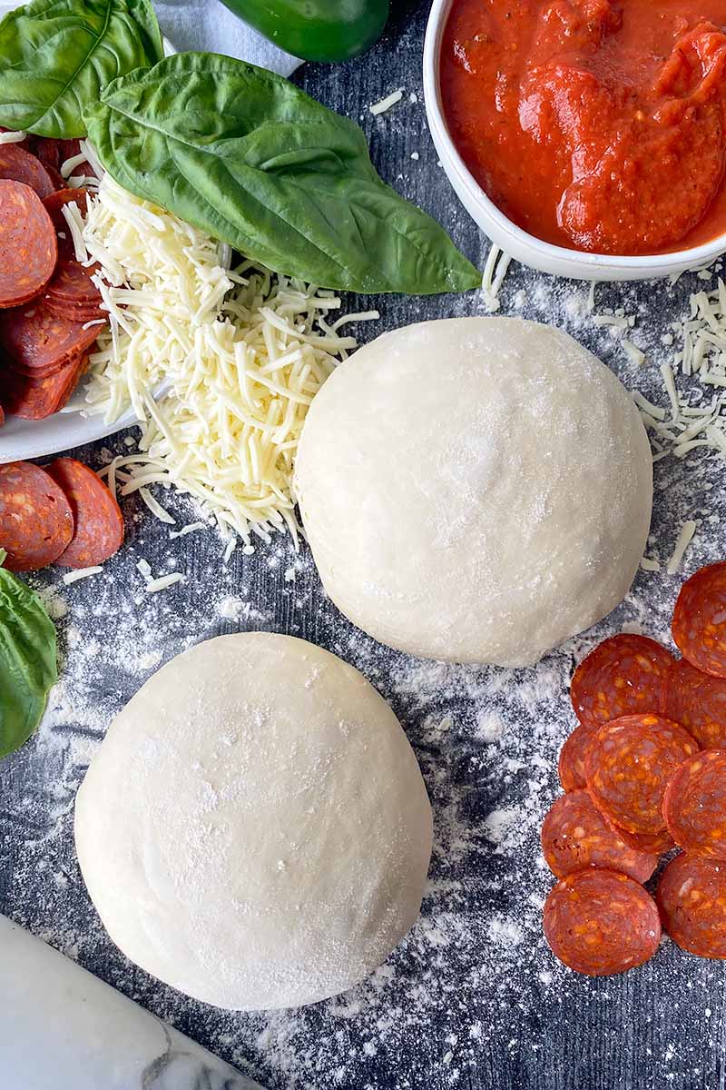 Vertical image of two mounds of lightly dusted unbaked bread on a floured surface next to cheese, basil, pepperoni slices, and a bowl of red sauce.