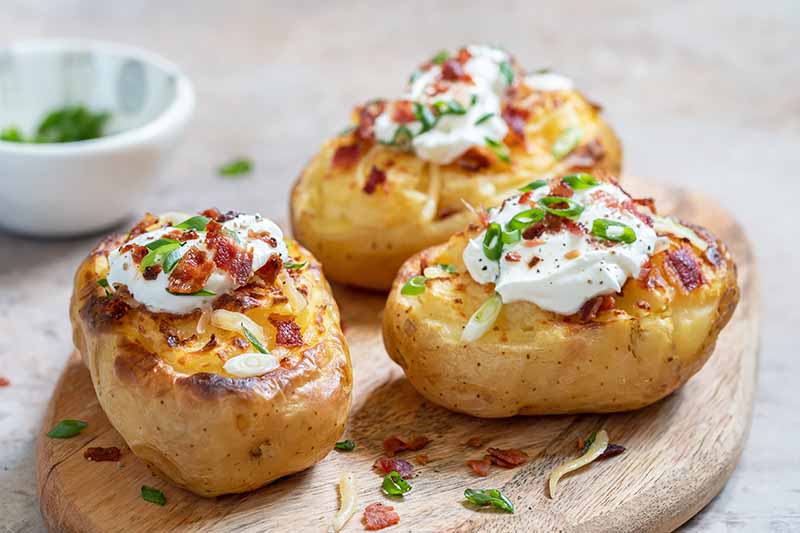 Horizontal image of three baked potatoes with bacon and chive garnishes and a dollop of dairy product on a plate.