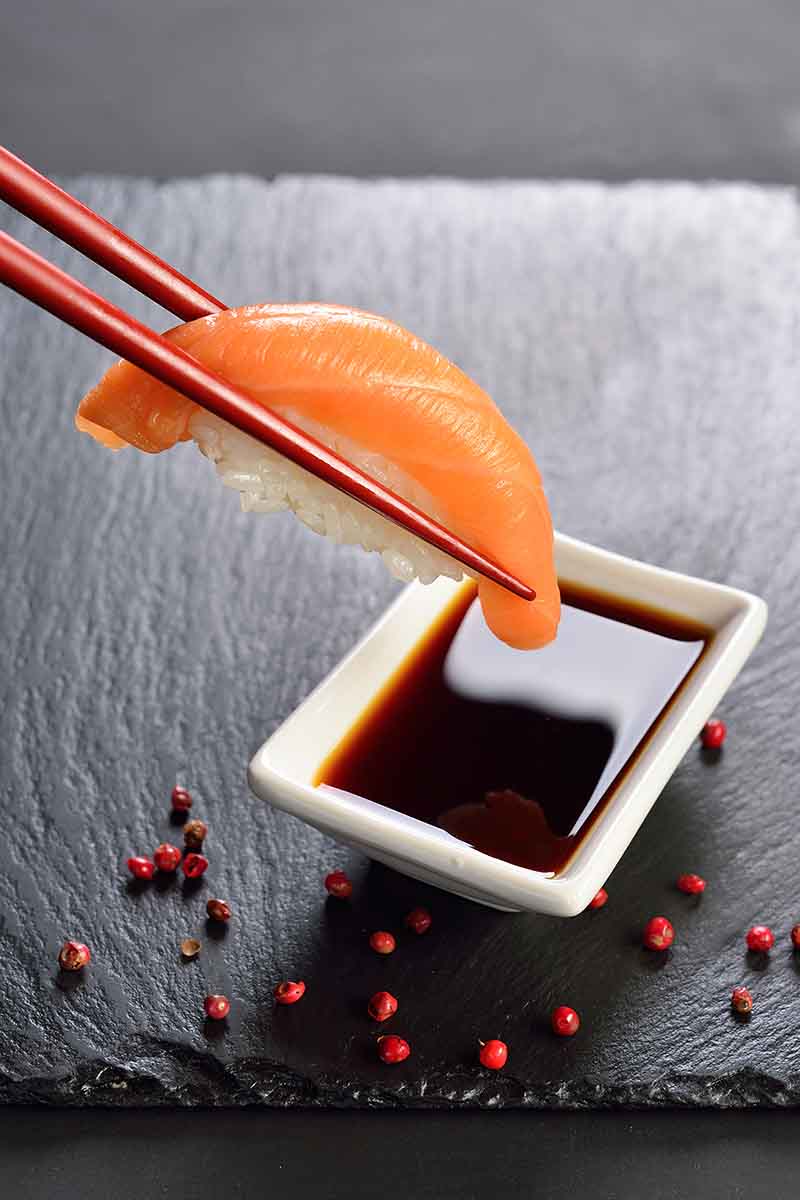 Vertical image of chopsticks dipping sushi into a bowl of dark brown liquid on a slate.