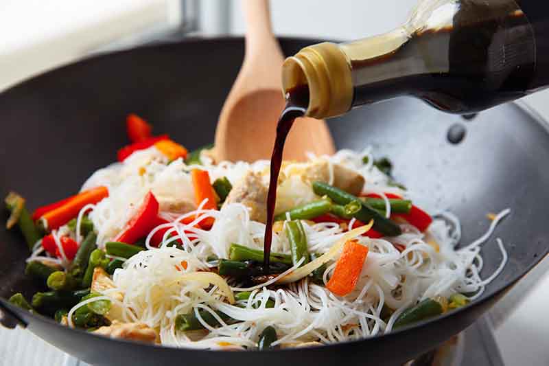 Horizontal image of pouring a dark brown liquid in a wok with noodles and vegetables.