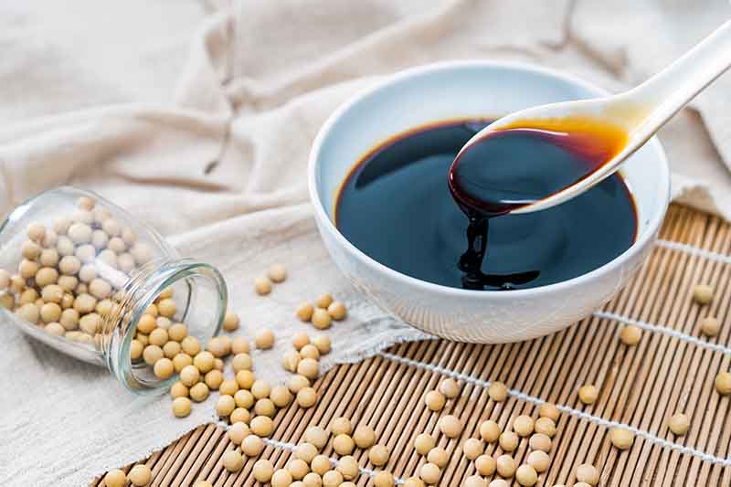 Horizontal image of a spoon lifting a dark brown liquid from a bowl next to a jar of spilled beans on a towel and bamboo mat.