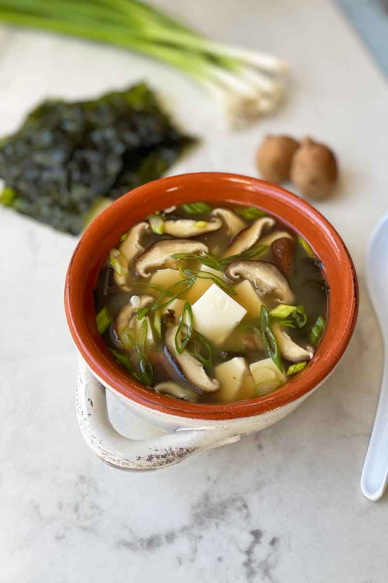 Vertical image of a red bowl with white handles filled with a broth with mushrooms and tofu cubes next to seaweed and scallions.