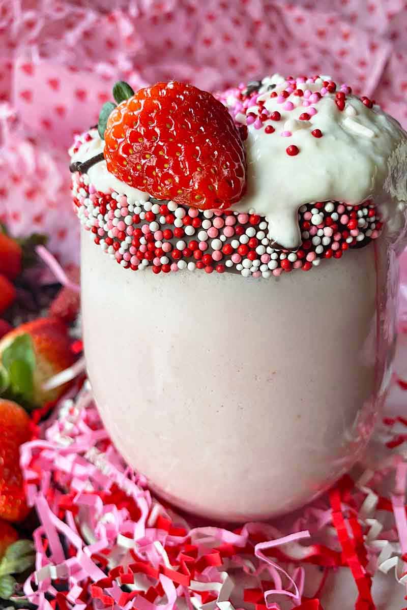 Vertical image of a glass cup filled with a light pink beverage garnished with fresh fruit, whipped cream, and sprinkles.