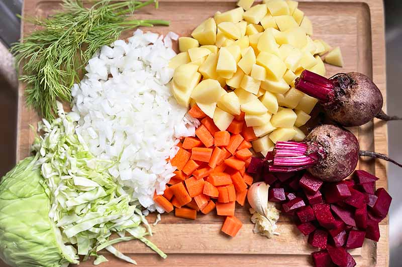 Horizontal image of assorted chopped vegetables and herbs on a wooden cutting board.