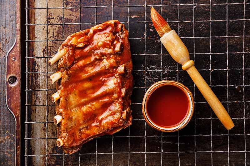 Horizontal image of a small rack of bone-in meat covered in red sauce next to a brush and a bowl of sauce on a baking sheet with a cooling rack.