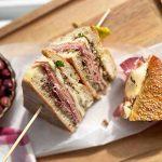 Horizontal image of a sliced cold cuts sandwich on parchment paper next to a bowl of kalamata olives.