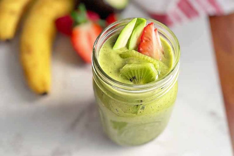 Horizontal image of a glass mason jar filled with a bright green mixture on a white surface next to bananas and strawberries.