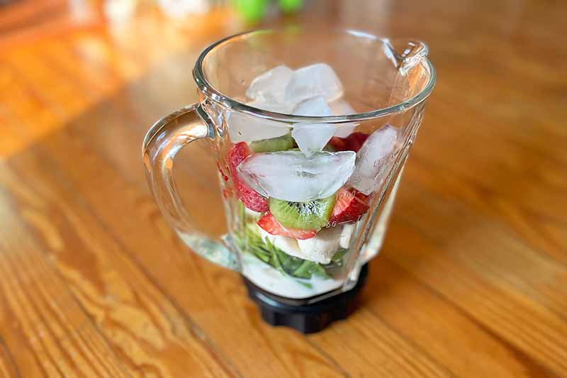 Horizontal image of a blender filled with fresh fruits and ice cubes on top on a wooden table.