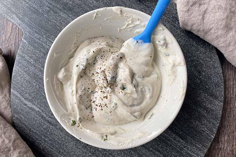 Horizontal image of mixing yogurt and spices together in a white bowl with a blue spatula.