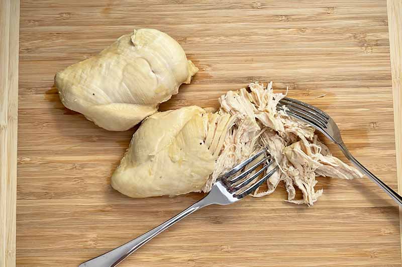 Horizontal image of shredding poultry breasts on a wooden cutting board with metal forks.