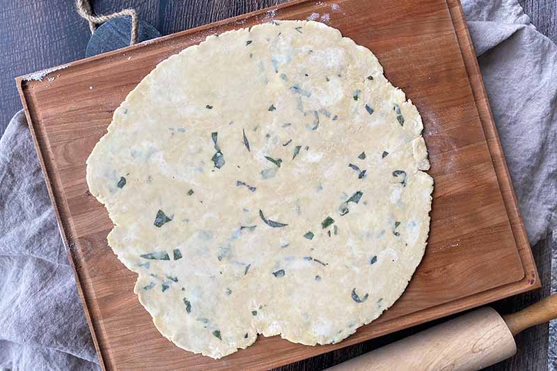 Horizontal image of a rolled out round dough speckled with chopped herbs on a wooden board.
