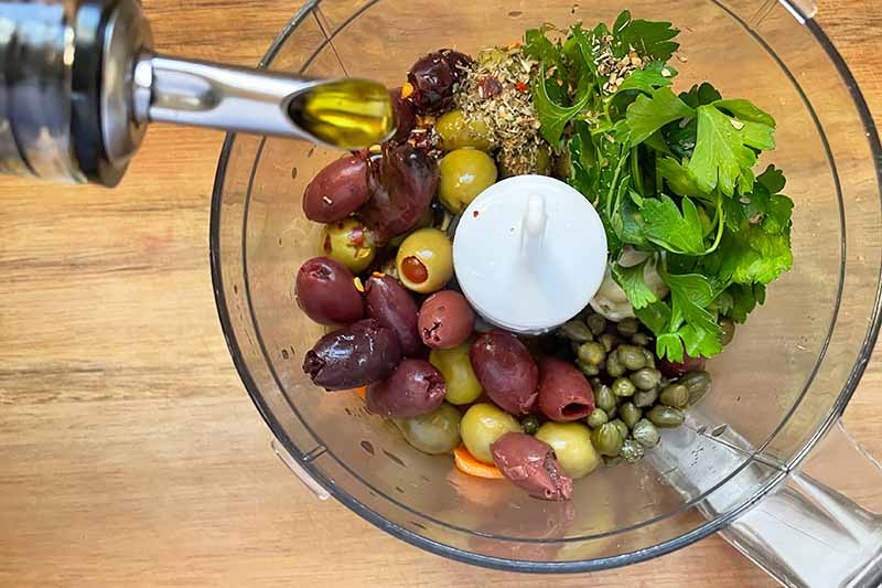 Horizontal image of pouring olive oil into a food processor with olives, capers, and herbs.