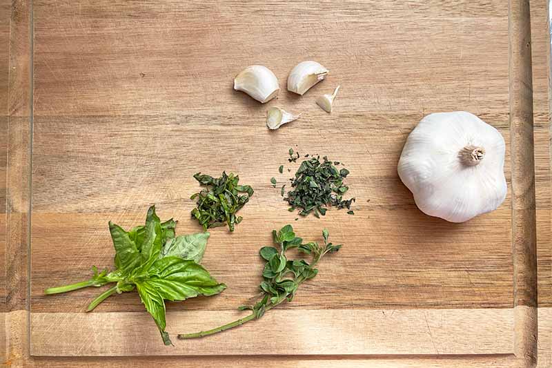 Horizontal image of prepped herbs and garlic on a wooden cutting board.