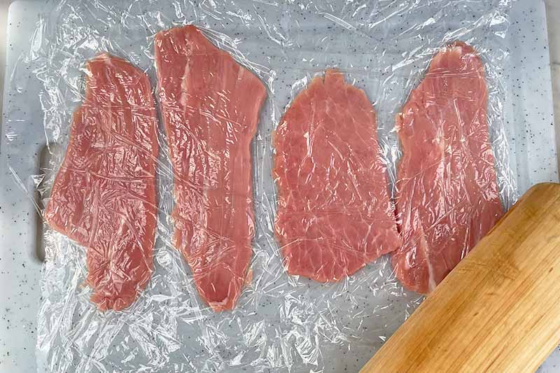 Horizontal image of pounded slices of raw meat between plastic wrap.