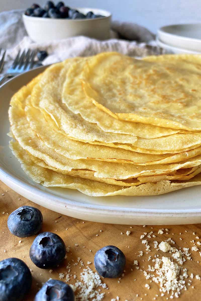 Vertical image of a pile of thin pancakes on a white plate next to fresh blueberries.