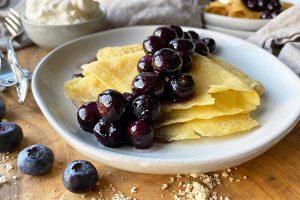 Horizontal image of a white plate with two folded crepes topped with blueberries on a wooden table in front of a bowl of whipped cream.