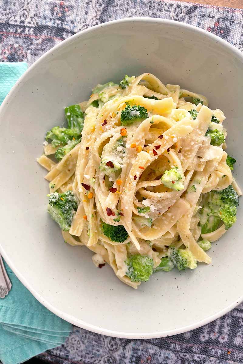 Vertical top-down image of a white dish filled with fettuccine in a creamy sauce with green vegetables garnished with red pepper flakes.