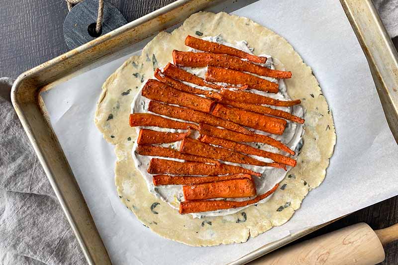 Horizontal image of a rolled out round dough with a white sauce and shingled carrots.