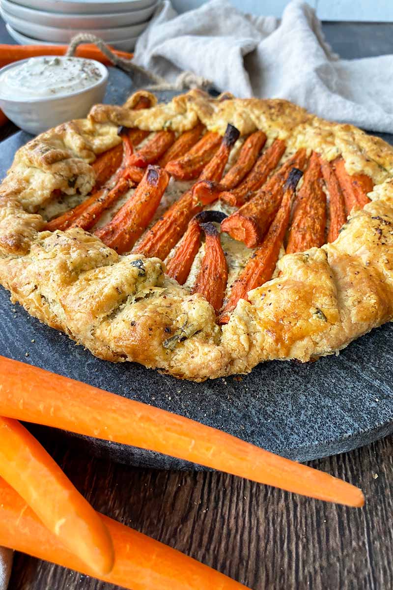 Vertical image of a round savory pastry with roasted orange vegetables on a slate next to more fresh vegetables, a bowl of dip, and a towel.