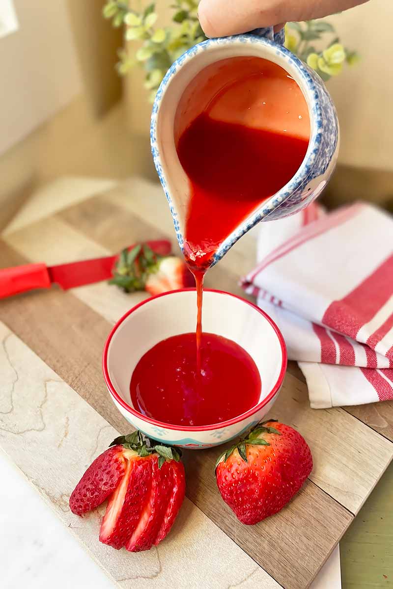 Vertical image of pouring a bright red sauce into a bowl surrounded by red fruit.
