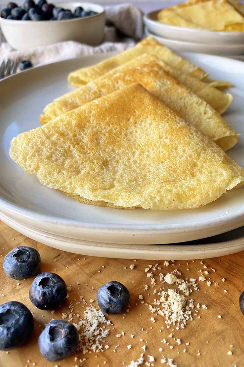 Vertical image of folded flat pancakes on a white plate next to fresh blueberries on a wooden surface.