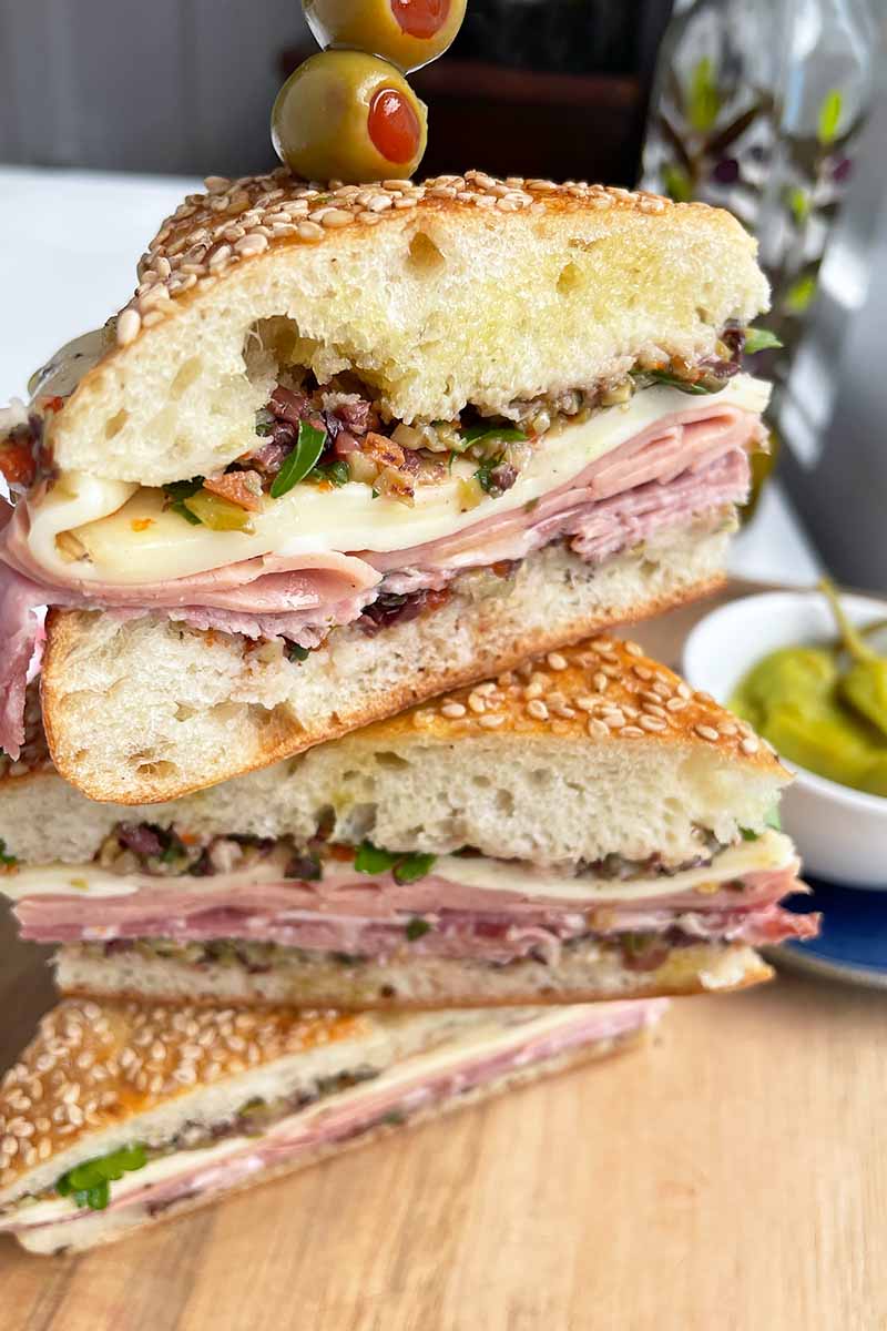 Vertical close-up image of a stacked muffuletta on a wooden cutting board with olives on a toothpick.