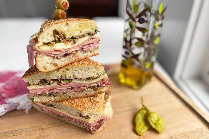 Horizontal image of a stack of sliced muffuletta on a wooden cutting board next to peppers and a glass of oil.