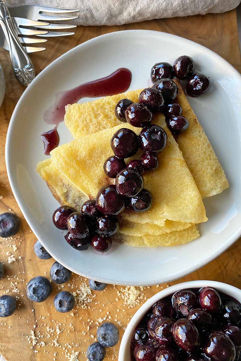 Vertical top-down image of two folded crepes on a white plate topped with a blueberry sauce.