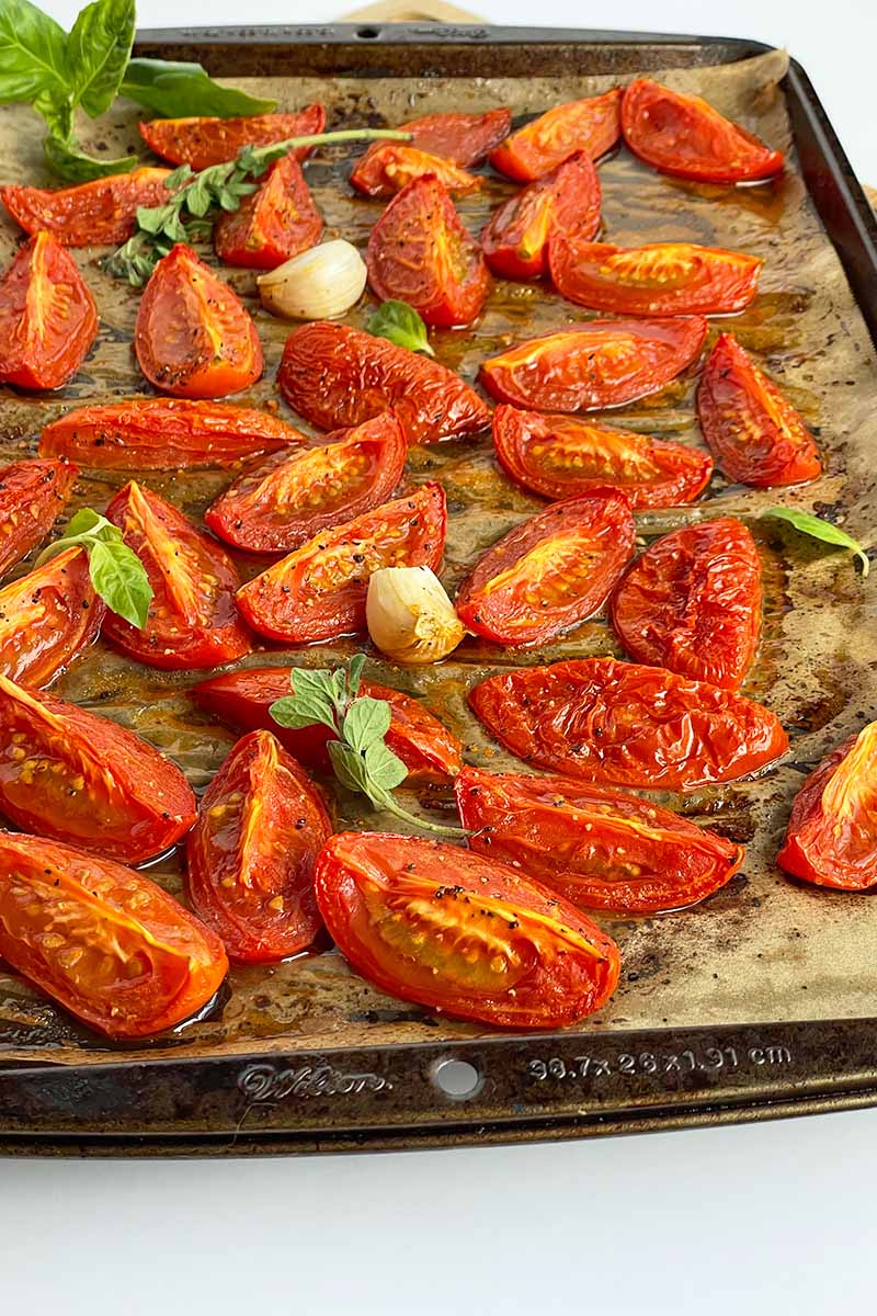 Vertical image of roasted slices of tomatoes, herbs, and garlic on a baking sheet.