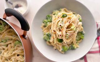 Horizontal image of fettuccine in a cream sauce with green vegetables in a white bowl next to a copper pot, bowl of grated cheese, and a red and white striped towel.
