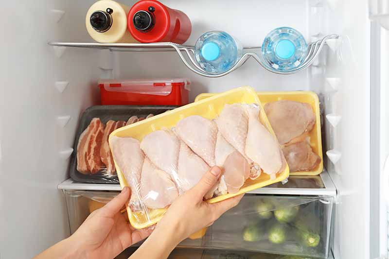 Horizontal image of packaged chicken legs held by two hands in front of an open refrigerator.