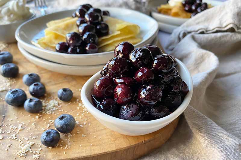 Horizontal image of a small white bowl with blueberries and a stack of white plates with folded crepes topped with more fruit.