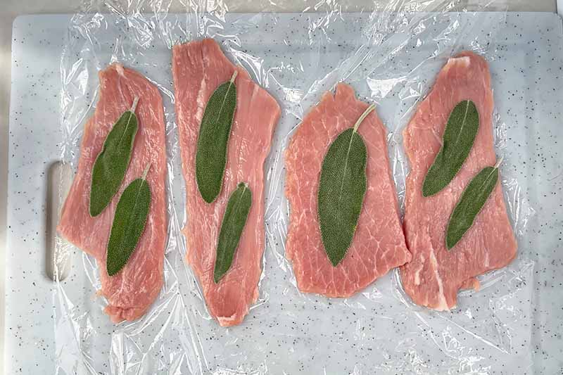 Horizontal image of layering fresh herbs on pounded slices of thin meat on plastic wrap.