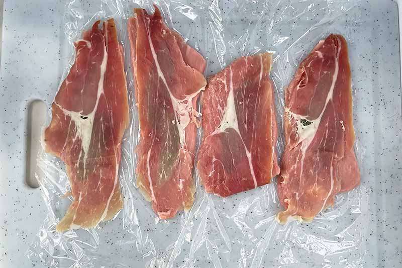 Horizontal image of prosciutto slices over slices of meat on plastic wrap.