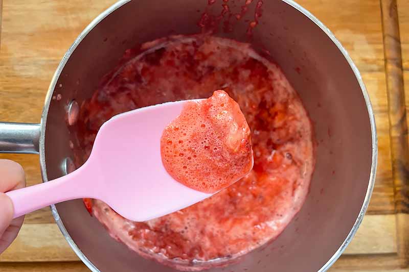 Horizontal image of removing foam with a pink spatula from a boiling red sauce.