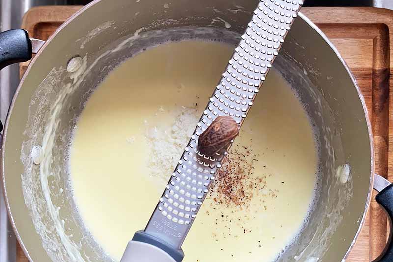 Horizontal image of grating nutmeg over a cream sauce in a pot.