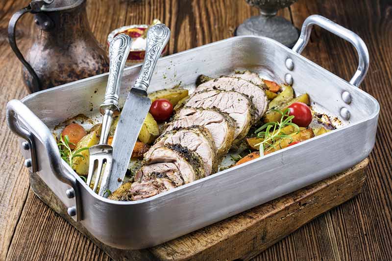 Horizontal image of a rectangular metal cookware with slices of cooked pork loin on top of vegetables and herbs with silverware on the side.