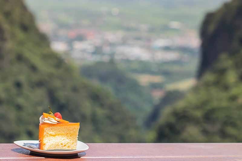 Horizontal image of a slice of orange cake on a white plate on a ledge overlooking a valley.