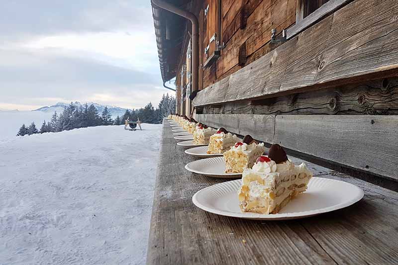 Horizontal image of slices of a dessert on whit plates on a wooden bench outside on a snowy ledge.
