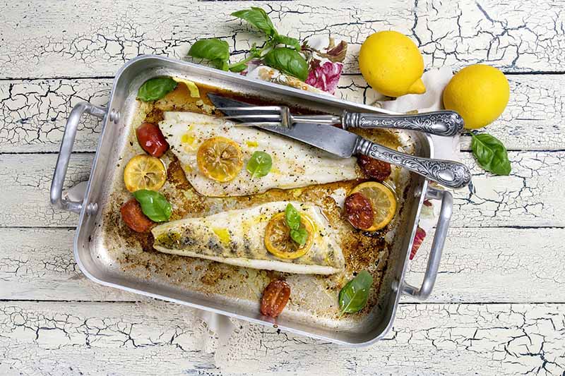 Horizontal top-down image of cooked fillets of fish with aromatics and lemon slices next to silverware.