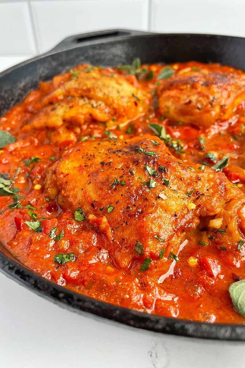 Vertical image of a cast iron skillet with pieces of poultry in a tomato sauce with fresh herbs on top.